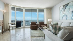 A modern living room with a large window offering a city view, a sofa, two armchairs, a coffee table, and a lamp. - Newly Remodeled Two Bedroom Condo with Immaculate Pool and Bay Views (Orange Beach)