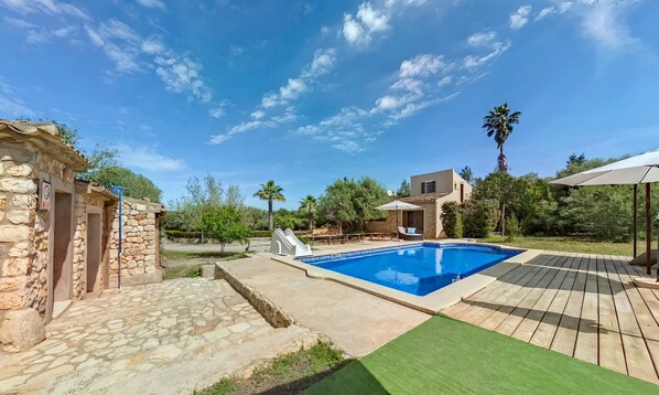 Une piscine avec une terrasse en pierre, un petit bâtiment aux murs de pierre et un palmier.