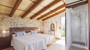 Une chambre avec un mur en pierre, des poutres apparentes, un lit double, une table de chevet, une commode, un miroir, une chaise et une douche.