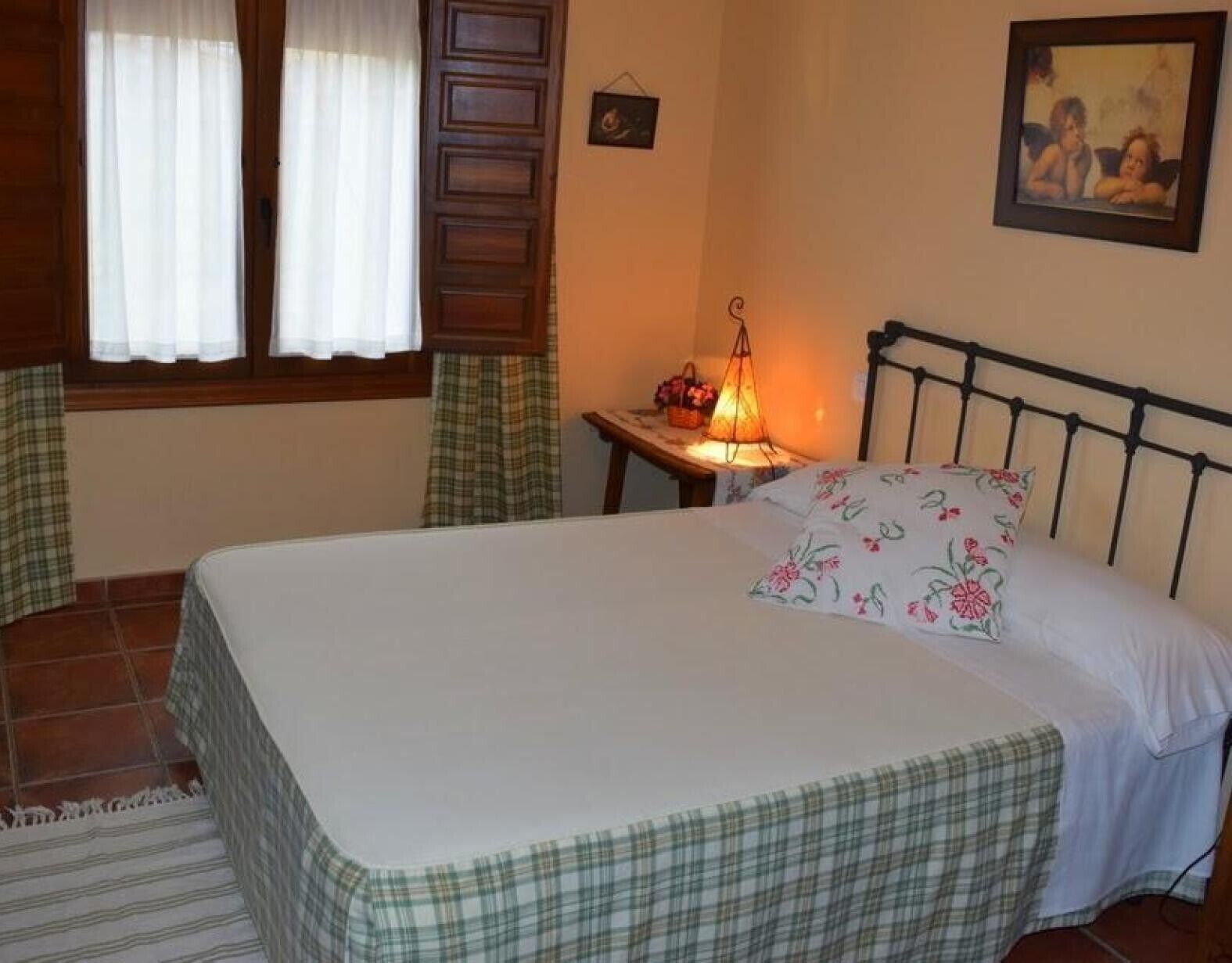 A bedroom with a bed, a nightstand with a lamp, a window with curtains, and a painting on the wall.