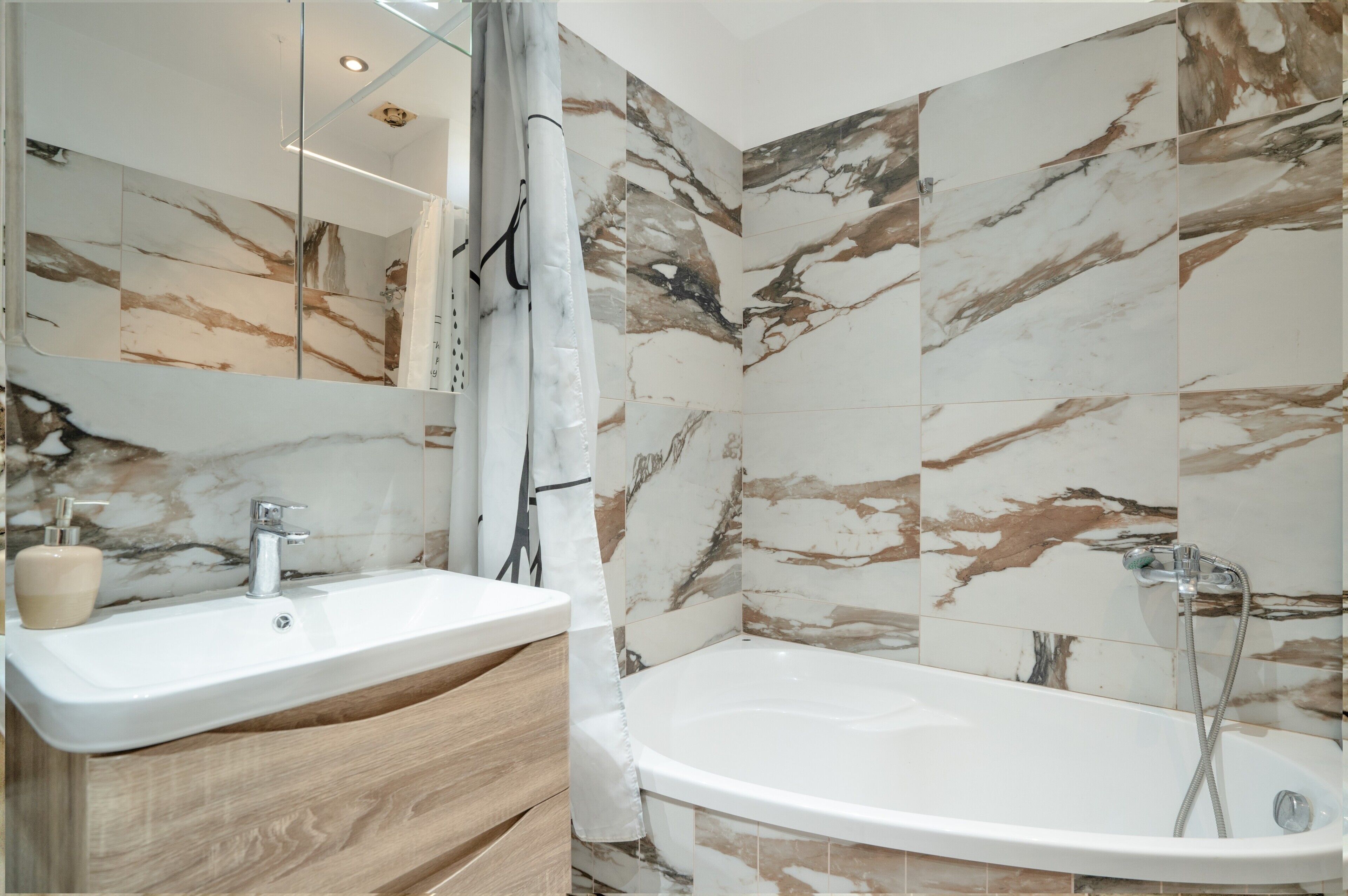 A bathroom with a marble-tiled wall, a white sink, a wooden vanity, and a freestanding bathtub.