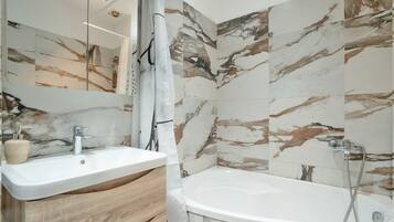 A bathroom with a marble-tiled wall, a white sink, a wooden vanity, and a freestanding bathtub.