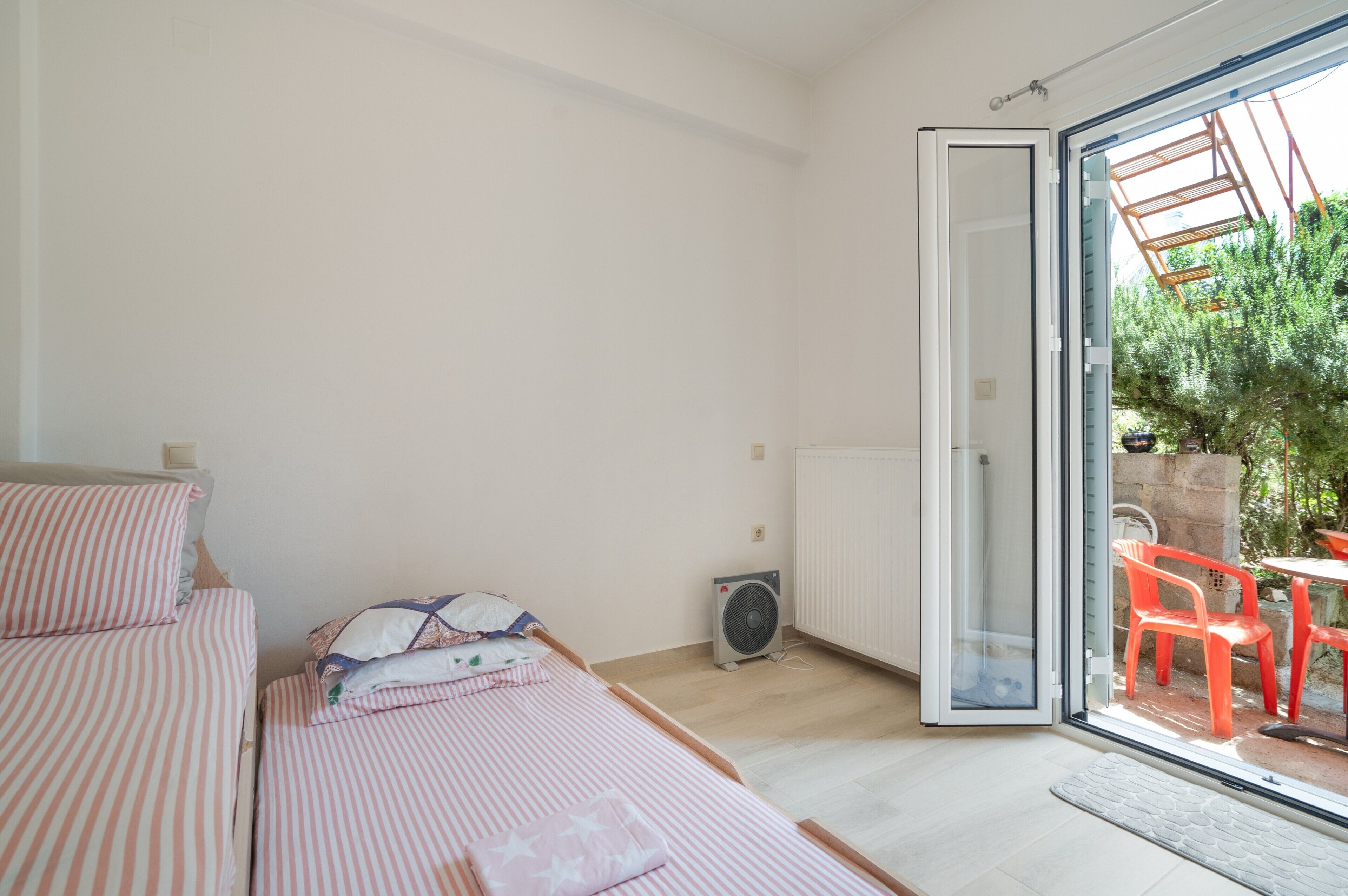 A bedroom with two beds, a radiator, an air conditioning unit, and a sliding glass door leading to an outdoor area with chairs and a table.