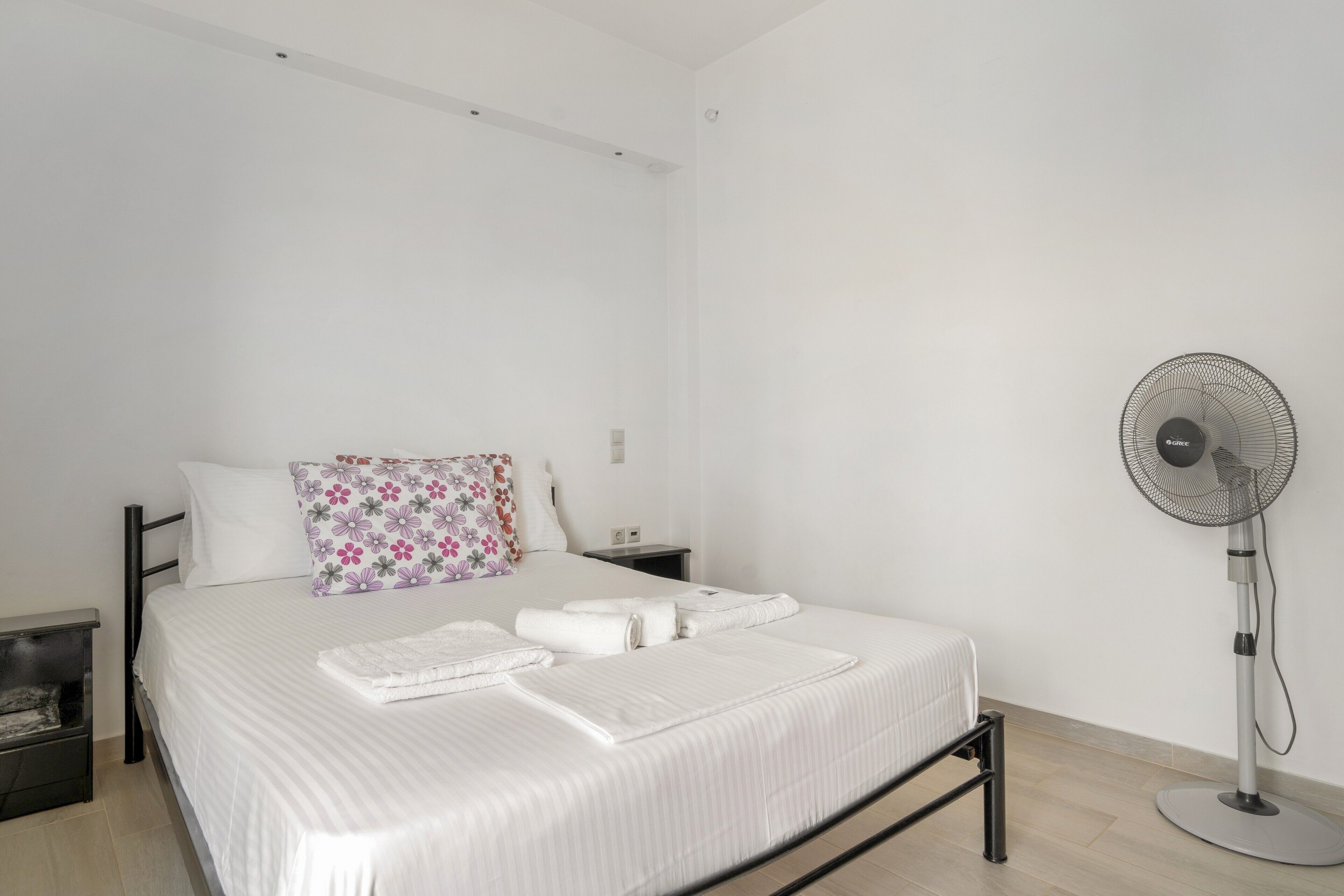 Ein Schlafzimmer mit einem Bett, Kissen, einem Nachttisch und einer Wandleuchte. Ein Standventilator ist ebenfalls vorhanden.
