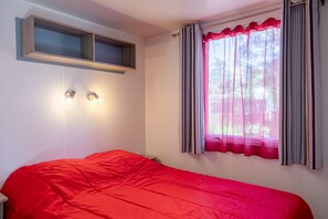 A bedroom with a single bed, a window with curtains, a shelf above the bed, and two wall-mounted lights. - Mobile Home on Family Campsite with Pools (Saint-Jean-de-Monts)