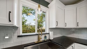 A kitchen with white cabinets, a black countertop, a stainless steel sink, and a window with a view of trees.