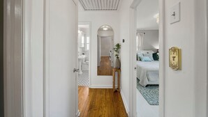 A narrow hallway with wooden flooring leading to a bedroom with a bed and a nightstand. A mirror reflects the room, and a door leads to another room.