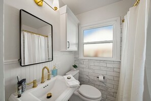 A bathroom with a white sink, gold faucets, a white toilet, and a window with a view of the beach.