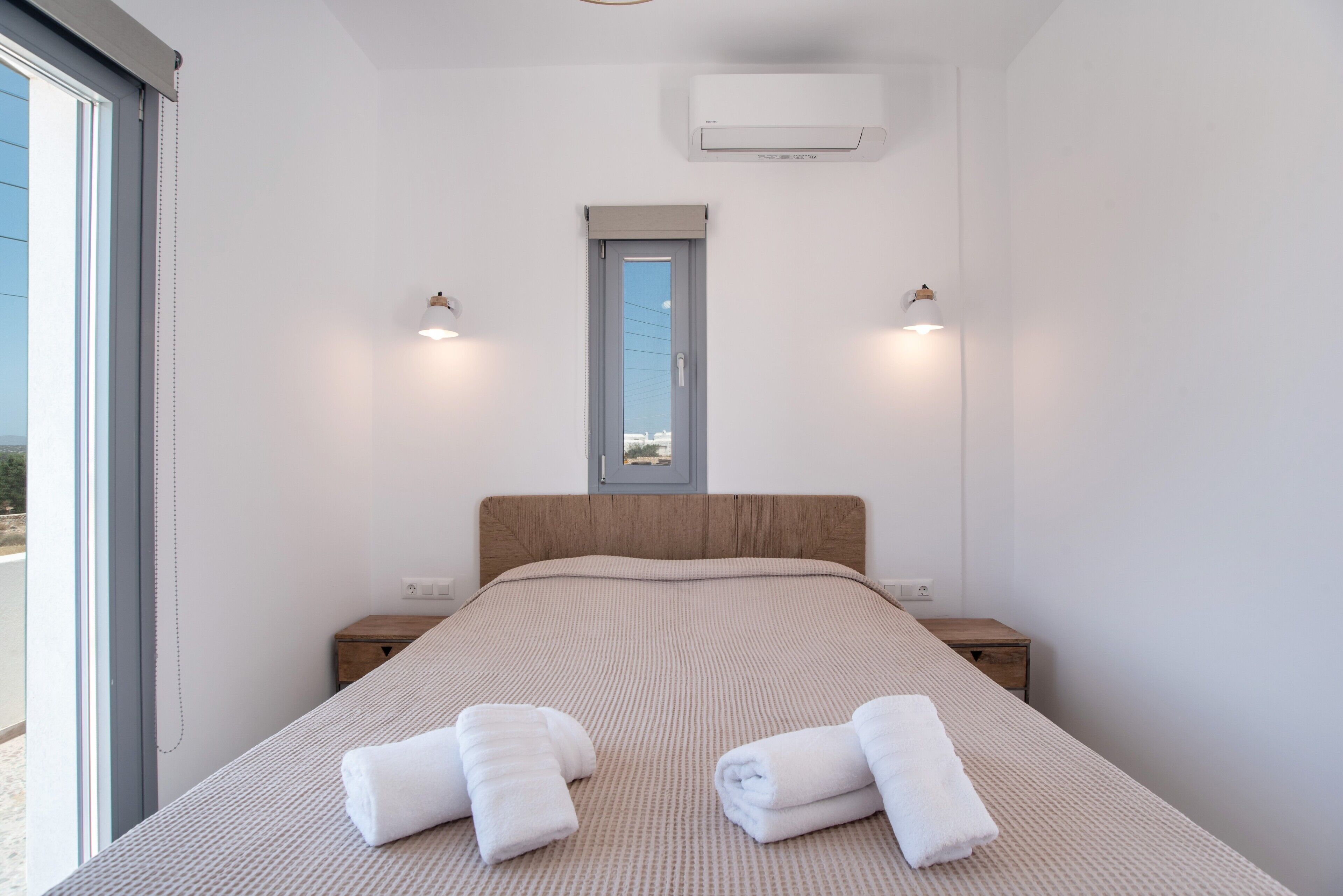 A bedroom with a bed, two bedside tables, a window with a view, and an air conditioning unit.