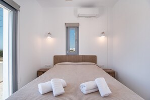 A bedroom with a bed, two bedside tables, a window with a view, and an air conditioning unit.