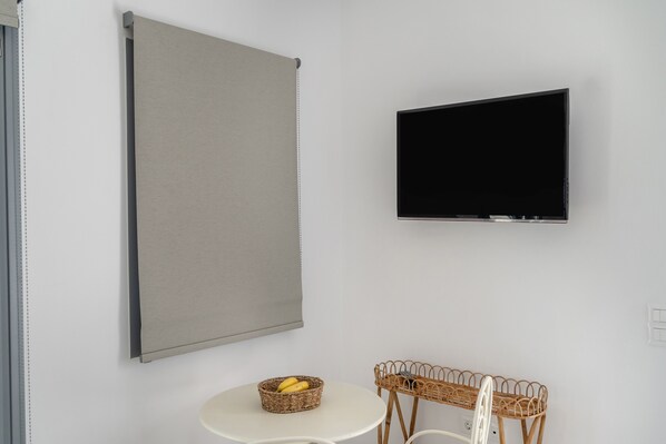 Una stanza con TV a parete, una grande tenda a rullo grigia, un tavolino rotondo con un cesto in vimini e un comodino in vimini con una sedia.