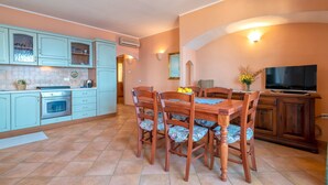 Een keuken met blauwgroene kastjes, een eettafel met bloemenstoelen, en een televisie aan de rechterkant.