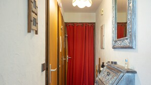 Un couloir étroit avec une porte en bois, un rideau rouge, un miroir et une commode surmontée d’un élément décoratif.