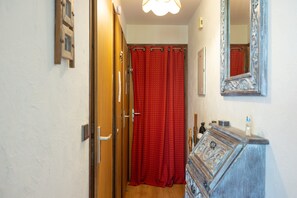Un couloir étroit avec une porte en bois, un rideau rouge, un miroir et une commode surmontée d’un élément décoratif.