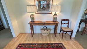 Una habitación con una consola de madera, dos lámparas, una silla, un tapete y un cuadro enmarcado en la pared.