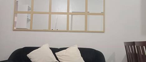 Quarto com sofá de cor escura, duas almofadas brancas e uma cadeira de madeira. Um espelho grande, parcialmente obscurecido, está pendurado na parede.