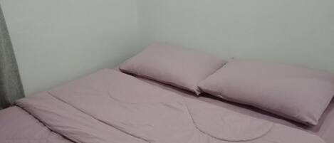 A single bed with a pink bedspread and pillows, a rolled white towel on the bed, and a plain wall.