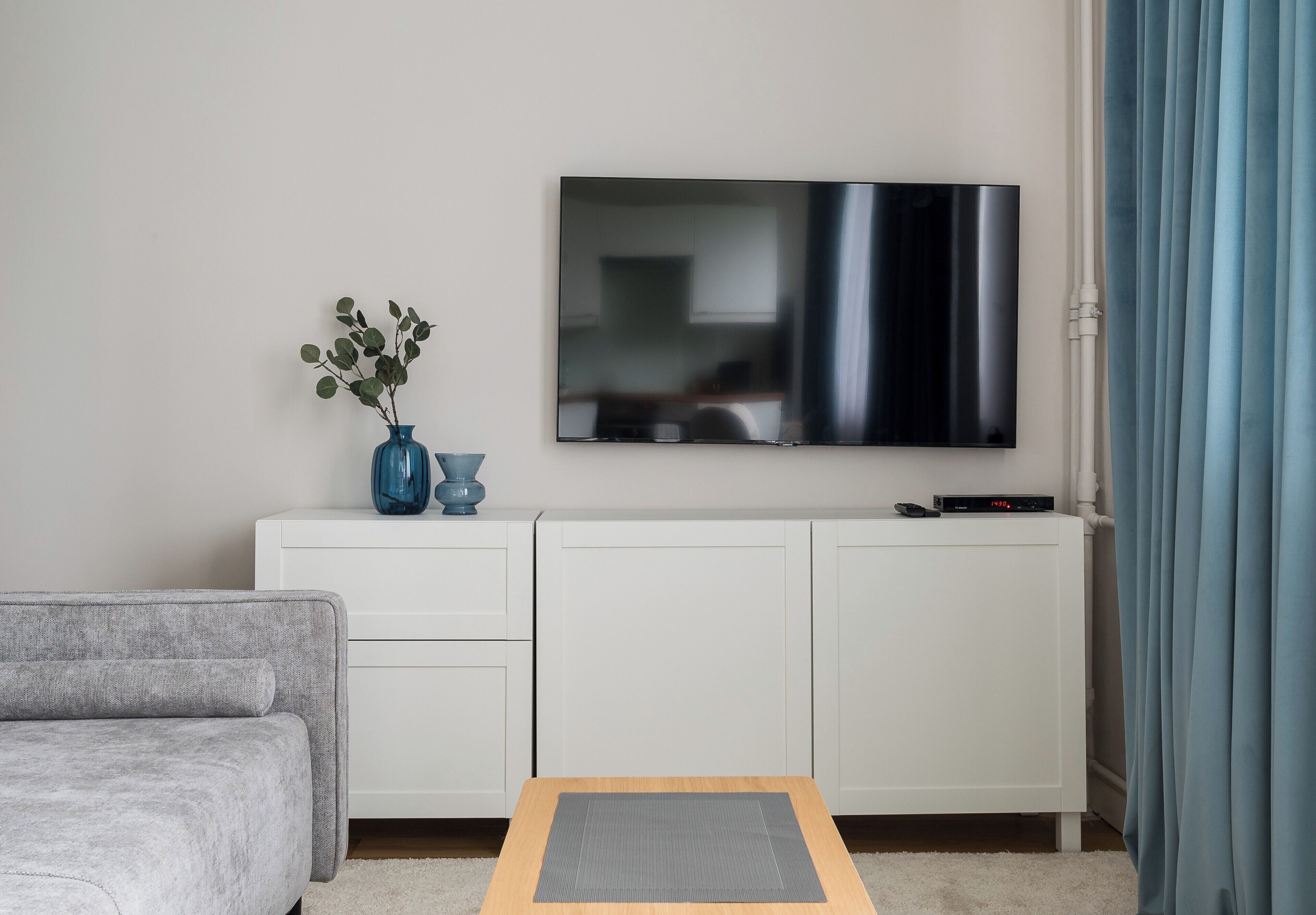 Una sala moderna con un televisor de pantalla plana montado en la pared, un sofá gris, un armario blanco y una pequeña mesa con un tapete gris.