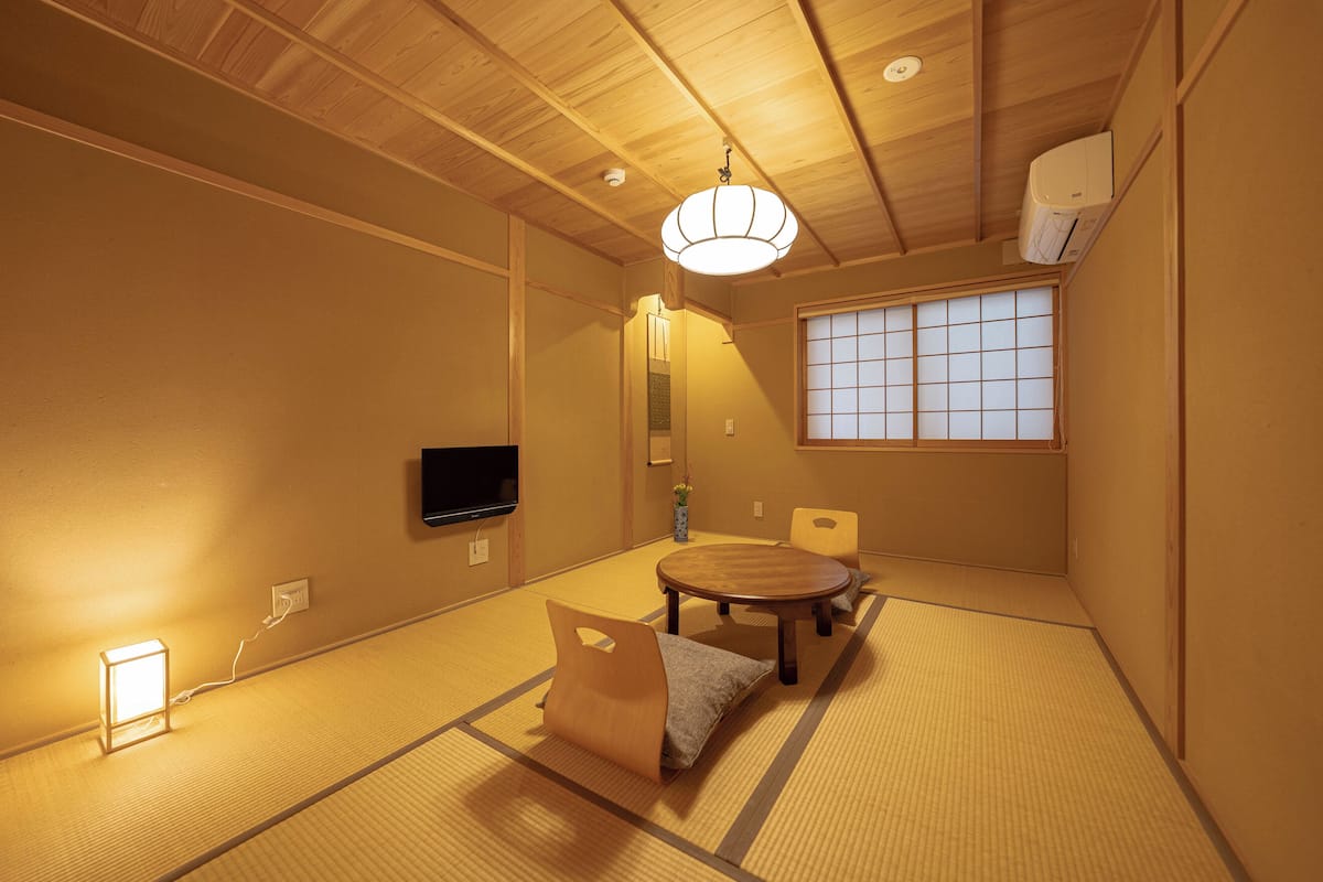 A traditional Japanese room with tatami flooring, a low table with cushions, a television, and a shoji screen window.