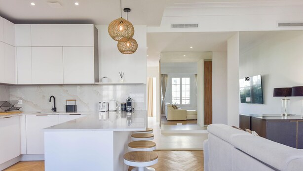 A modern kitchen with white cabinetry, a central island with stools, a hanging light fixture, and a view into a living area with a sofa and a TV.