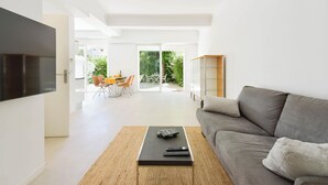 Un salon moderne avec un canapé gris, une table basse et un téléviseur fixé au mur. On aperçoit un coin repas avec des chaises et une table à travers une porte vitrée.