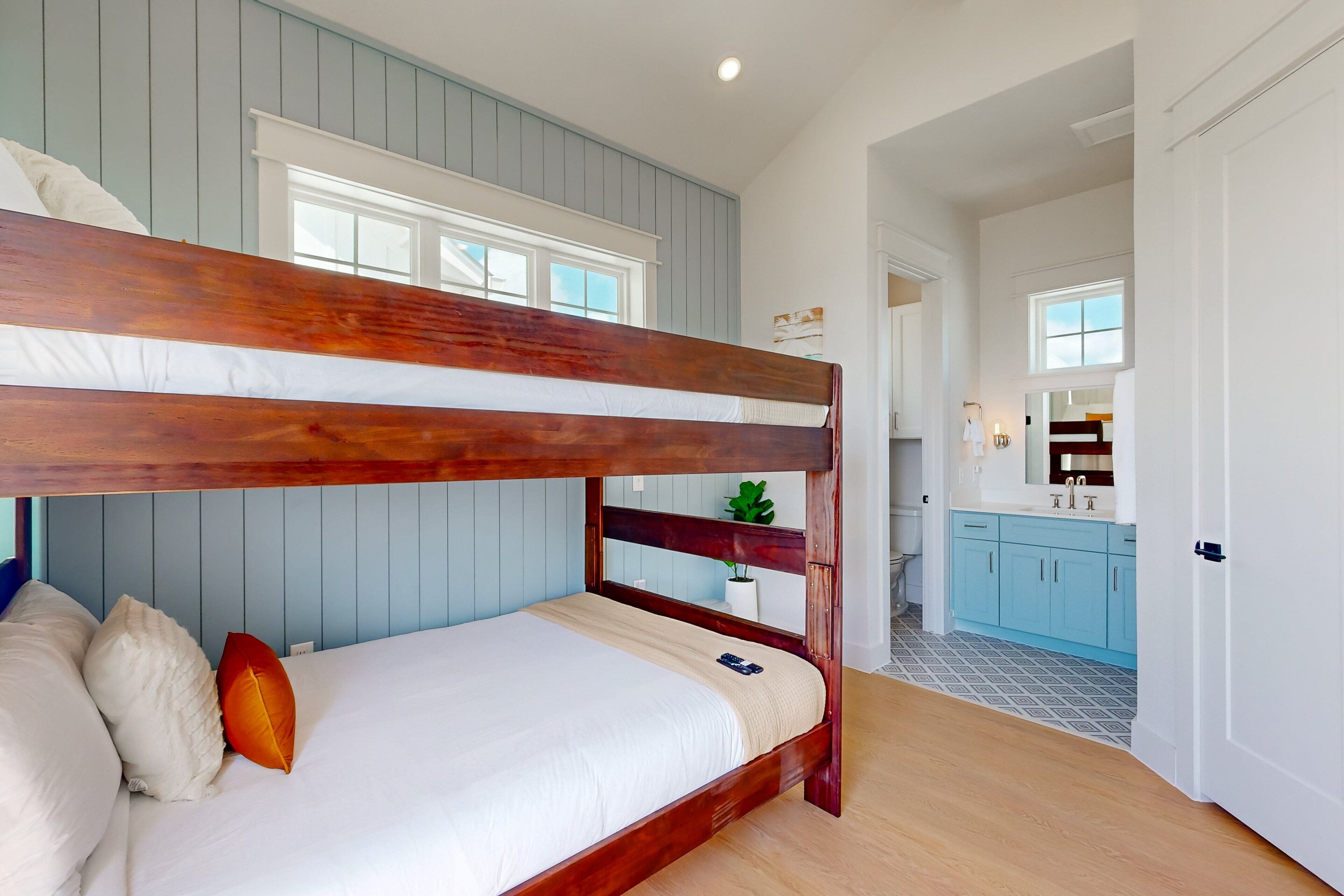 A bunk bed with white bedding and a wooden headboard, a potted plant, and a kitchen area with blue cabinets and a white countertop.