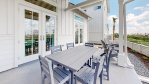 Un patio cubierto con una mesa de comedor y bancos, ventanas grandes y vistas a la playa.