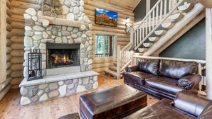 A log cabin interior with a stone fireplace, leather sofa, and a staircase with wooden railings.