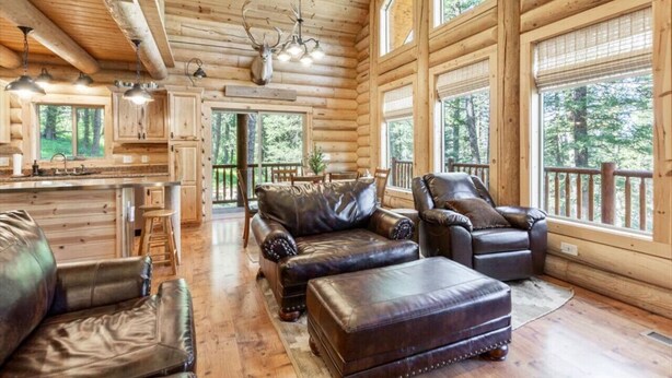 A log cabin interior with a central seating area, wooden walls, large windows, and a kitchen.