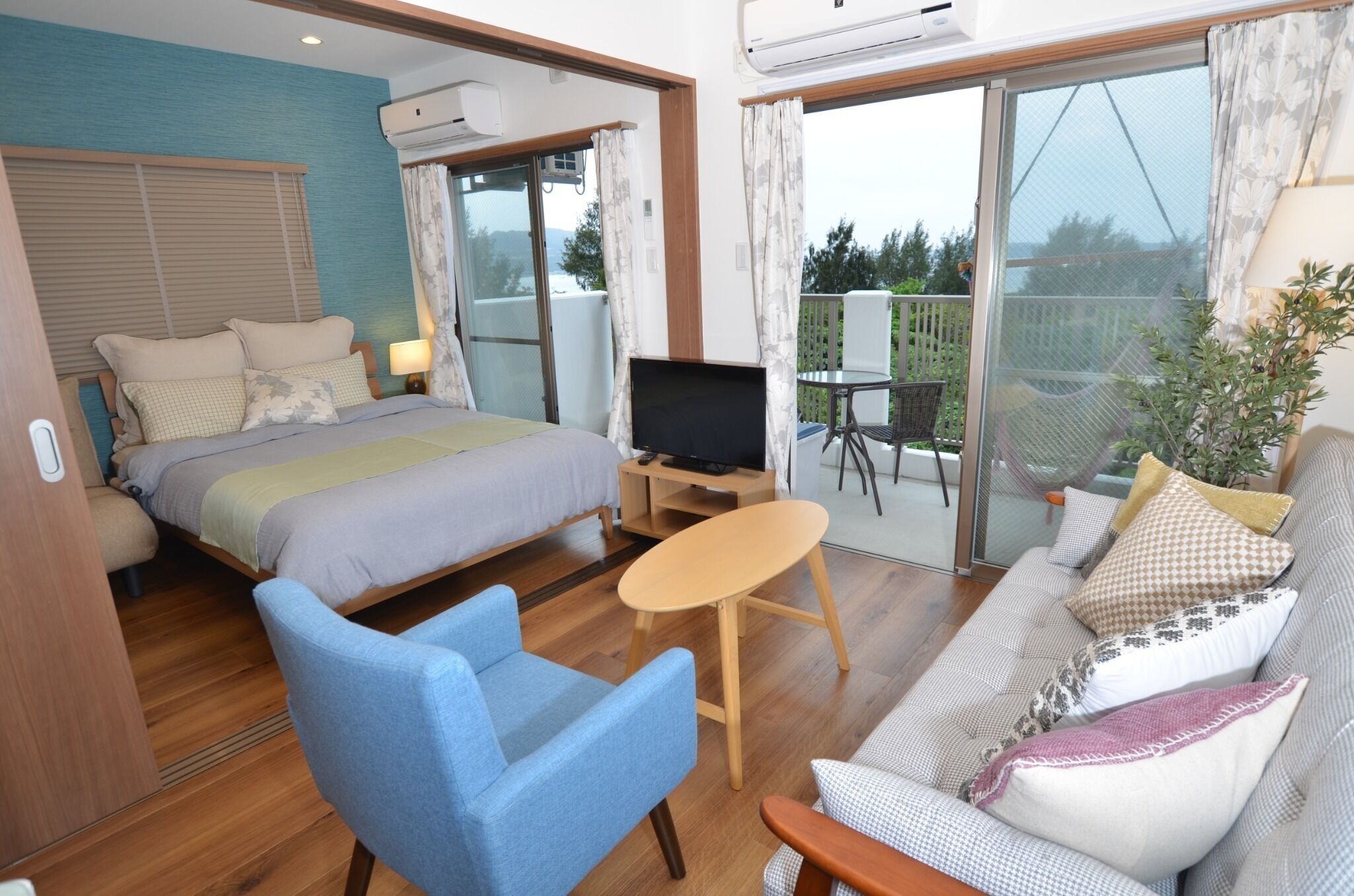 A room with a bed, a TV on a stand, a blue chair, a sofa, and a balcony with a view.