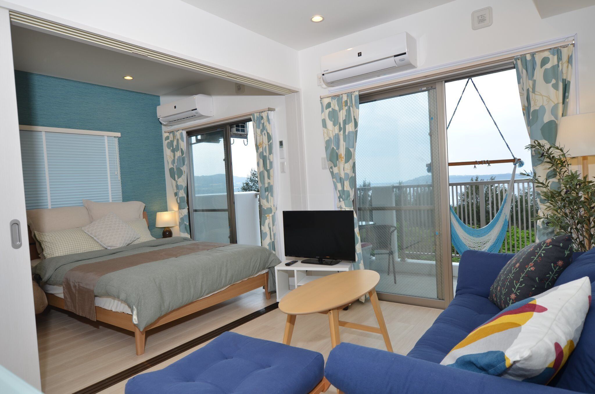 A bedroom with a bed, a TV, a small table, a sofa, and a balcony with a hammock.
