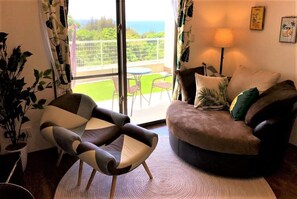 Een woonkamer met een bank, fauteuil en uitzicht op een balkon met een tafel en stoelen.