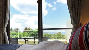 Habitación de hotel con un ventanal grande, cama con ropa blanca, almohada a rayas y vista a zonas verdes y una cerca en el exterior.