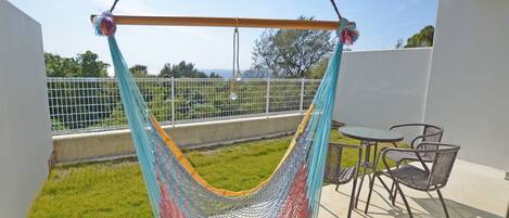 Una hamaca colgada de una viga de madera en un balcón, con vistas a un área de césped y árboles.