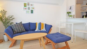 Una sala moderna con un sofá azul, una mesa de centro de madera y un reposapiés a juego. Hay una mesa de comedor blanca con sillas, una zona de cocina y una planta en maceta.