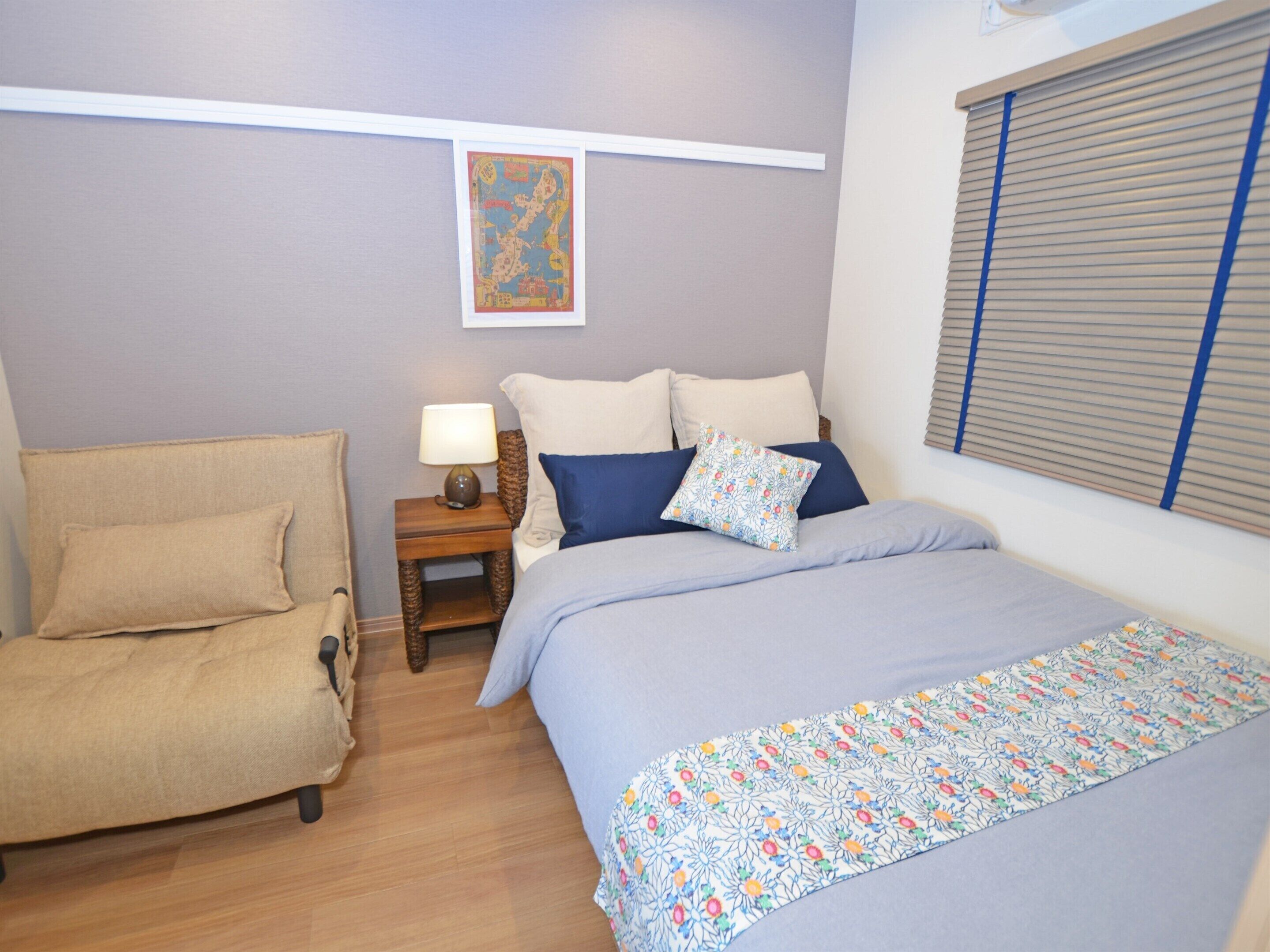 Un dormitorio con cama, una silla, una mesita de noche con lámpara y una ventana con persianas.