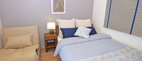 Un dormitorio con cama, una silla, una mesita de noche con lámpara y una ventana con persianas.