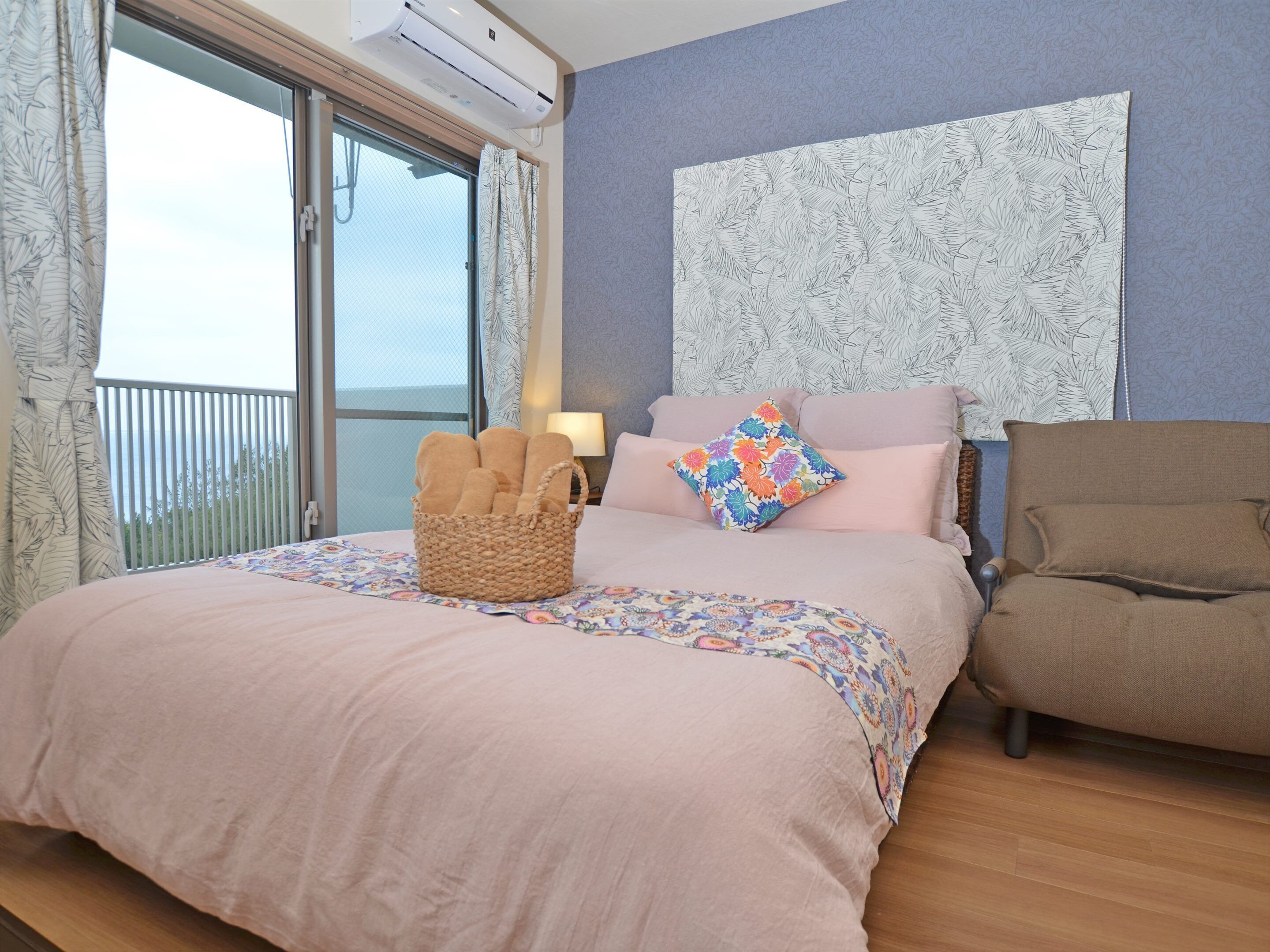 Un dormitorio con cama, una silla, una cesta de toallas, un ventanal y una pared con diseño de hojas.
