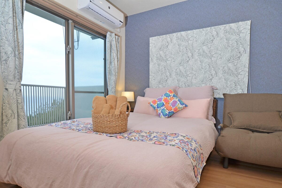 A bedroom with a bed, a chair, a basket of towels, a window, and a wall with a leaf pattern.