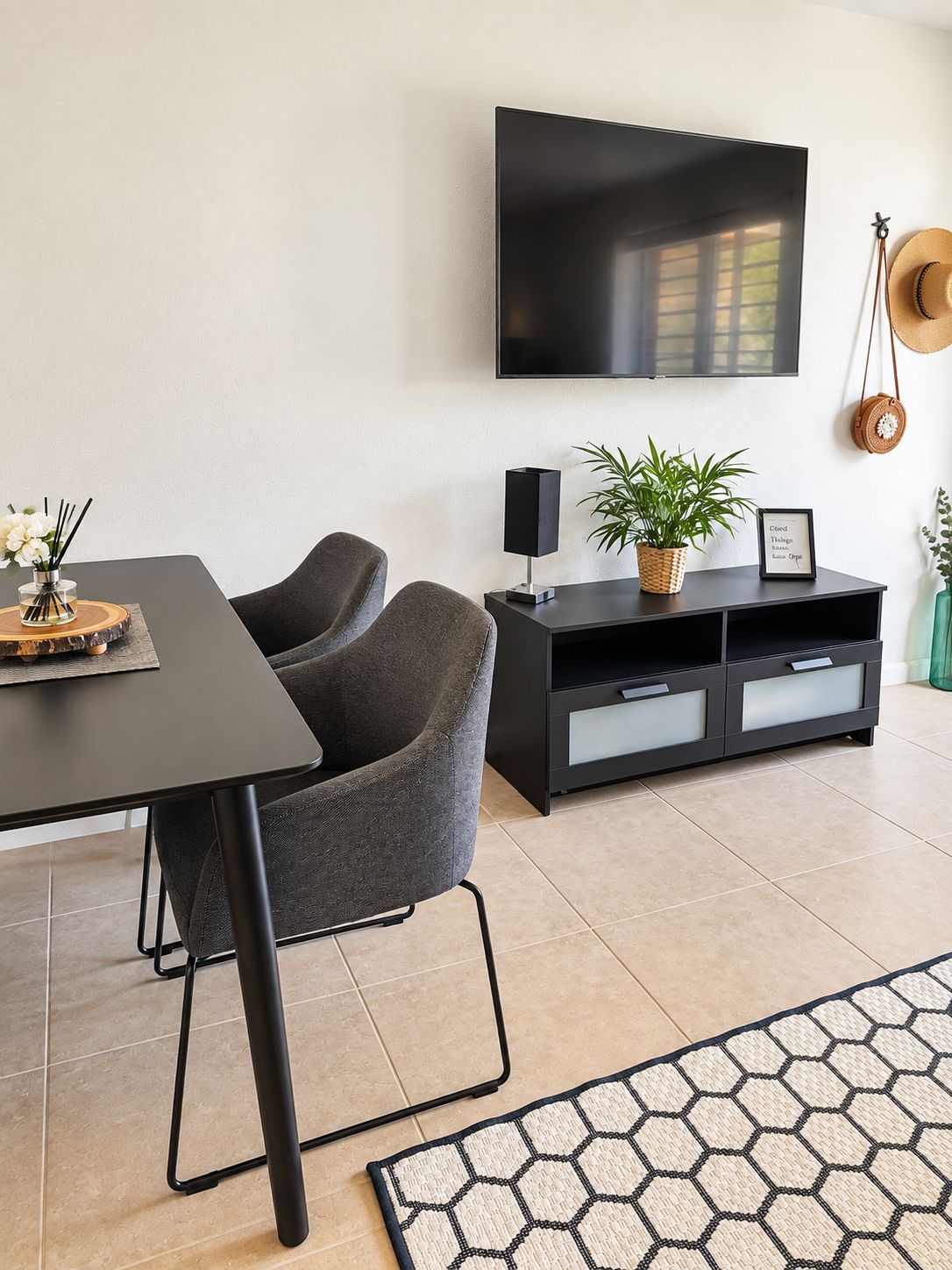 A modern living room with a flat-screen TV mounted on the wall, a dark wooden dining table with chairs, a black entertainment unit with a plant, and a hexagonal patterned rug.