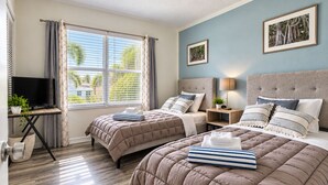 A hotel room with two beds, a TV, a window with curtains, a bedside table with a lamp, and a wall-mounted picture. - Florida Beach Hideaway 2A– Near the Beach • Pool • Fast WiFi • free Parking (Fort Myers)