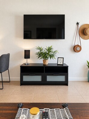 Un salon moderne avec un téléviseur à écran plat fixé au mur, un meuble noir pour appareils électroniques agrémenté d’une plante et d’un tableau encadré, un fauteuil gris, une lampe et un panier en osier.