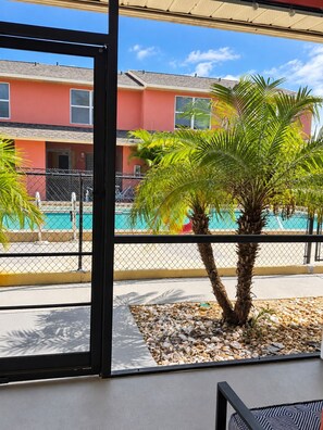 On aperçoit, à travers une porte vitrée, un espace piscine bordé de palmiers, avec un bâtiment en arrière-plan.