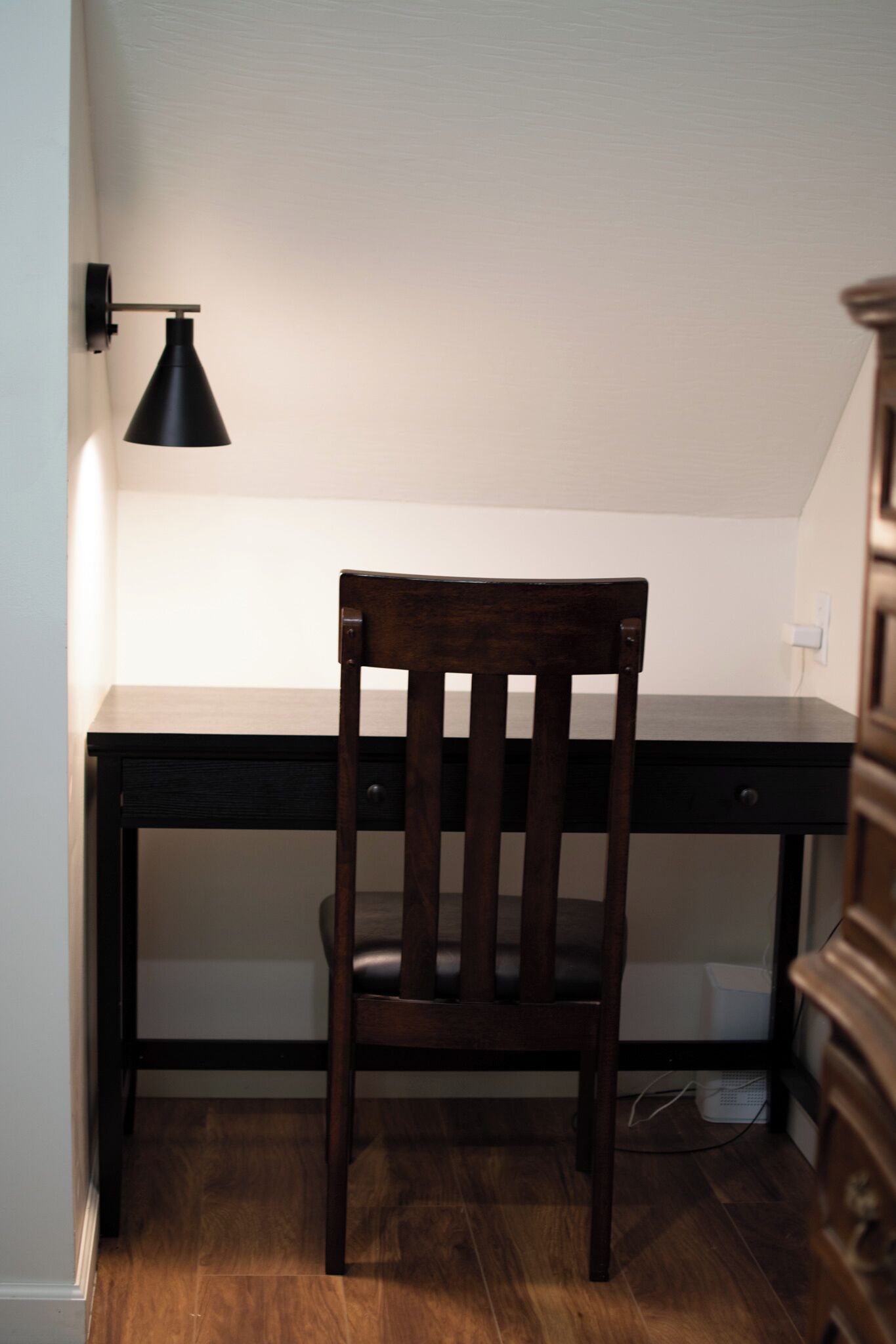 A wooden desk with a chair, a black wall lamp, and a white wall socket.