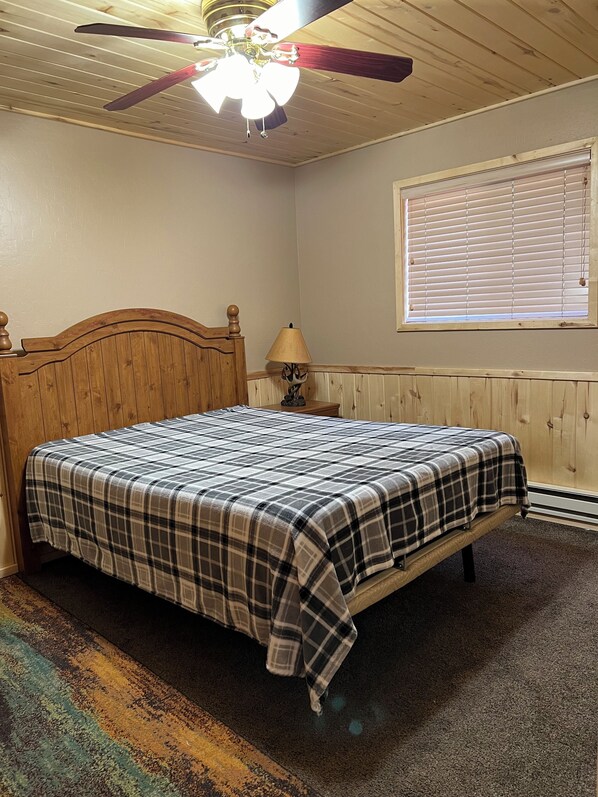 A bedroom with a wooden bed, a ceiling fan, a window with blinds, and a lamp on a nightstand. - Life is better in a cabin. (Heber-Overgaard)