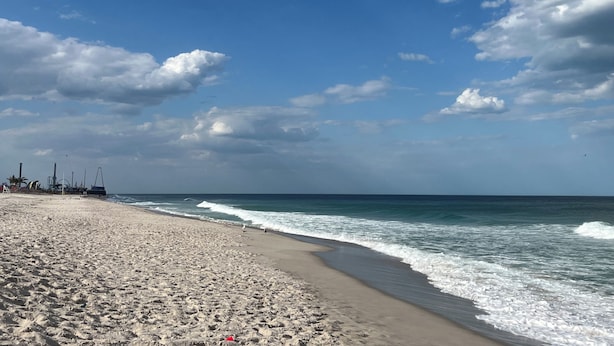 Une vaste plage de sable fin, avec des vagues douces, un ciel dégagé et, au loin, des bâtiments industriels.