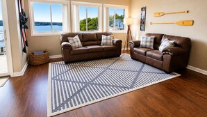 Sala de estar con dos sofás de cuero marrón, alfombra estampada, piso de madera y vista a un lago a través de ventanas grandes.