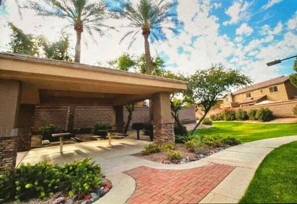 A covered patio area with stone pillars, a brick pathway, and a lawn with palm trees.