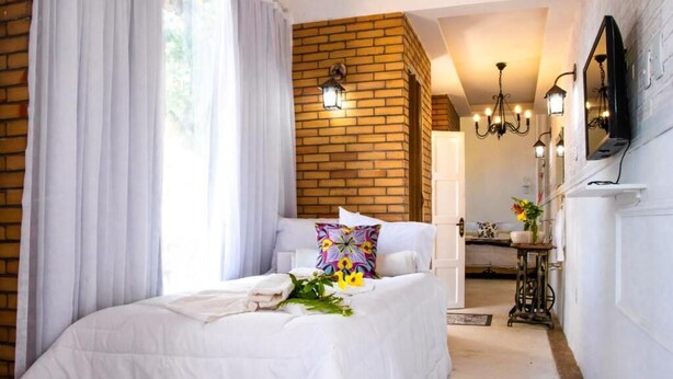 A bedroom with a bed, white curtains, a brick wall, a chandelier, and a television on the wall. - Pousada Saruê Natal (Natal)