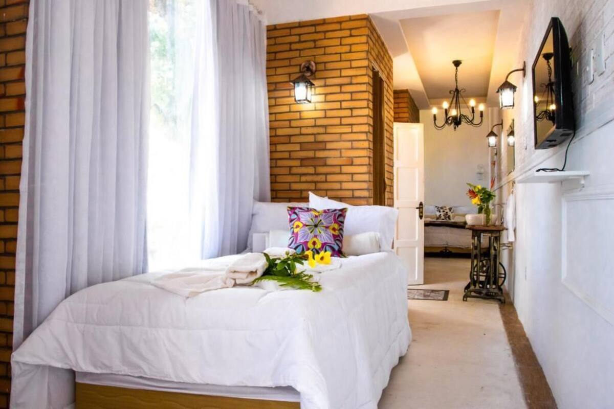A bedroom with a bed, white curtains, a brick wall, a chandelier, and a television on the wall.